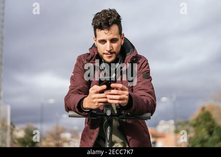 Schöner Kerl sendet eine Nachricht mit seinem Handy auf dem Elektroroller. Konzentrieren Sie sich auf den Mann. Stockfoto
