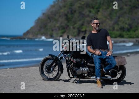Rüde mit Bart und Sonnenbrille auf seinem Motorrad Am Strand Stockfoto