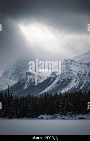 Ein Loch in den Wolken lässt Lichtstrahlen über den schneebedeckten kanadischen Rockies, wie man es vom verschneiten Herbert Lake im Banff National Park aus sieht. Stockfoto