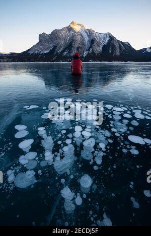 Ein Abenteurer sitzt vor einem abstrakten Array der gefrorenen Eisblasen des Abraham Lake, wenn die Sonne untergeht an dem Tag. Stockfoto
