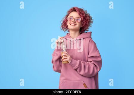 Lächelndes Mädchen mit rosa Haaren in rosa Hoodie und Sonnenbrille trinken Obst Smoothie oder Saft aus Glasflasche auf blauem Hintergrund. Gesunde Ernährung, Gemüse Stockfoto