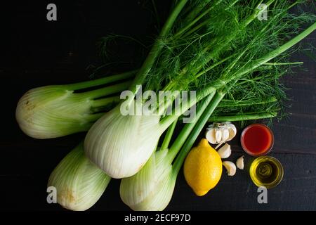 Sautierte Fenchel mit Knoblauch und Tomatensauce Zutaten: Rohe Fenchelzwiebeln, Zitrone, Knoblauch, natives Olivenöl extra und Tomatensaft auf dunklem Holz Stockfoto