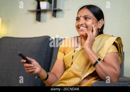 Indische Frau in Saree Fernsehen zu Hause, während sie sitzt Auf dem Sofa durch Halten der Fernbedienung während der Freizeit Stockfoto