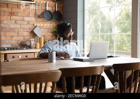 Selbstbewusster afroamerikanischer Mann, der in der Küche ein Headset trägt und einen Laptop verwendet Stockfoto