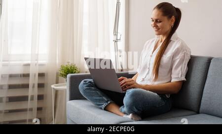 Lächelnde glückliche Frau, die auf dem Sofa sitzt und einen Laptop benutzt. Das Konzept der Remote-Arbeit oder Untersuchung während der Quarantäne. Stockfoto