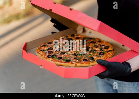 Lebensmittelkurier in schwarzen medizinischen Handschuhen hält rote Box mit Pizza von Pizzeria. Lebensmittellieferung bei Quarantäne Coronavirus covd-19 Zeitraum. Stockfoto