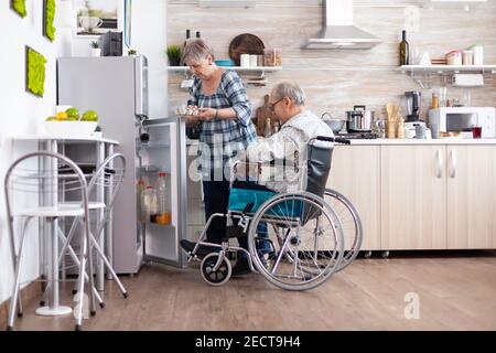 Ältere Frau bereitet Frühstück für behinderte Mann unter Eierkarton aus dem Kühlschrank, leben mit Mann mit Gehbehinderungen. Behinderter älterer Mann im Rollstuhl, der seiner Frau in der Küche hilft. Stockfoto