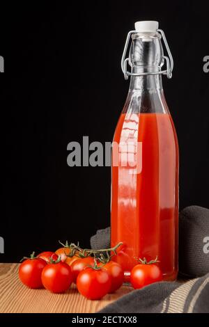 Ansicht der alten Glasflasche mit Tomatensaft, auf Holz mit Kirschtomaten, dunkles Tuch, schwarzer Hintergrund, vertikal Stockfoto