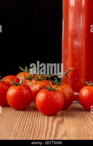 Nahaufnahme von Kirschtomaten auf Holz mit einer Flasche Tomatensaft, schwarzer Hintergrund, vertikal, mit Kopierraum Stockfoto