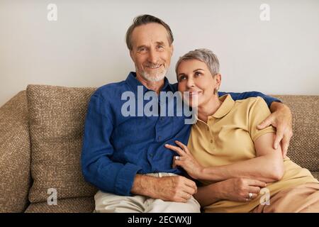 Porträt eines lächelnden älteren Ehepaares, das auf einer Couch im Wohnzimmer sitzt. Zwei positive reife Menschen zu Hause umarmen. Stockfoto