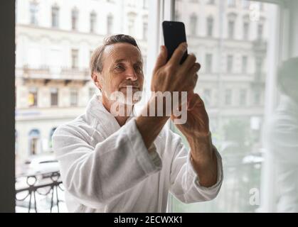 Ein älterer Mann im Bademantel steht auf dem Balkon eines Hotels Zimmer und ein Selfie machen Stockfoto
