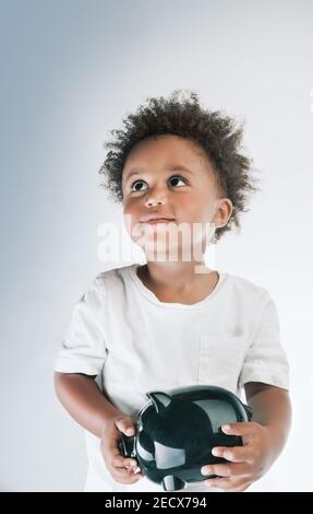 Porträt eines niedlichen kleinen afroamerikanischen Jungen Modell mit Piggy Bank in den Händen. Isoliert auf grauweißem Hintergrund. Investition in die Zukunft. Stockfoto