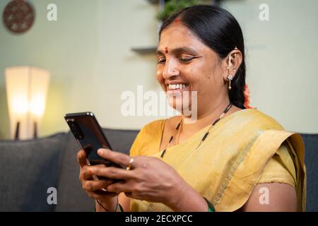 Glücklich lächelnde indische Frau beschäftigt mit Handy, während auf dem Sofa sitzen - Konzept der Menschen mit sozialen Medien, Online-Messaging-App, Internet und Stockfoto