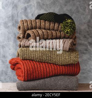 Stapel gefaltetes Strickpullover aus Wolle, Schal, Hut mit Bommel im Regal gegen graue Wand. Saison der warmen Strickmode Stockfoto