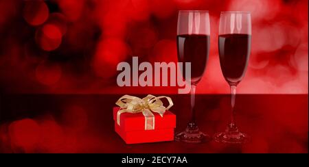 Zwei Weingläser und rote Geschenkbox auf dem Schreibtisch mit rotem Blur Bokeh Hintergrund, Sektgläser und Geschenke mit Feier und Jubiläum, Liebe und rom Stockfoto