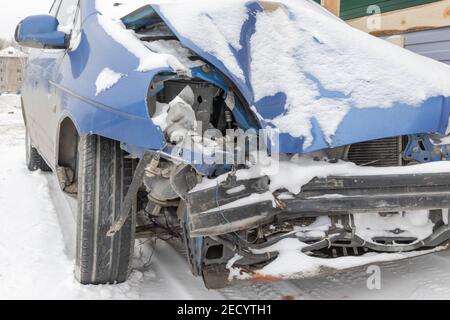 Blau abgestürzt Auto ohne Scheinwerfer und Stoßfänger nach einem Unfall Geparkt an der Straße unter einer Schneeschicht auf Ein kalter Wintertag Stockfoto