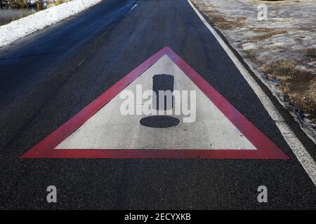 Nahaufnahme des roten weißen Dreiecks Warnschild Asphalt der rutschigen Straße mit Schnee und schwarzem Eis (Fokus auf der Mitte des Zeichens) Stockfoto