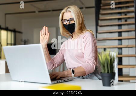 Elegante Frau mittleren Alters sitzt am Laptop im Büro während einer Pandemie, grüßt Kollegen, chattet mit Freunden, lächelt, gibt das hoch Stockfoto