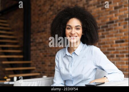 Erfolgreiche selbstbewusste junge Mixed-Race-Unternehmerin oder eine Geschäftsfrau mit Afro-Frisur in lässiger Kleidung, lächelnd und im Stuhl sitzend Stockfoto