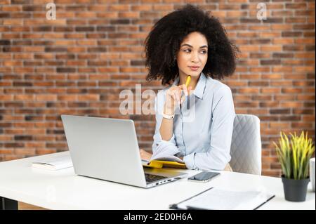 Nachdenkliche afroamerikanische Frau mit lockiger Frisur, die über die neue Idee nachdenkt, einen Stift hält, ihre Pläne überprüft, von zu Hause aus arbeitet, davon träumt Stockfoto