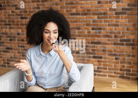Junge Geschäftsfrau mit gemischter Rasse oder eine Unternehmerin mit Afro-Frisur, die lässige Kleidung trägt, auf dem Stuhl sitzt und ein Mobiltelefon hält Stockfoto