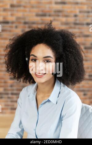 Junge multirassische Frau mit lockigen Frisur sieht direkt in die Kamera, Lächeln weit, hat eine Stirn Haut und weiße Zähne und ein leichtes Make-up, trägt ein Stockfoto