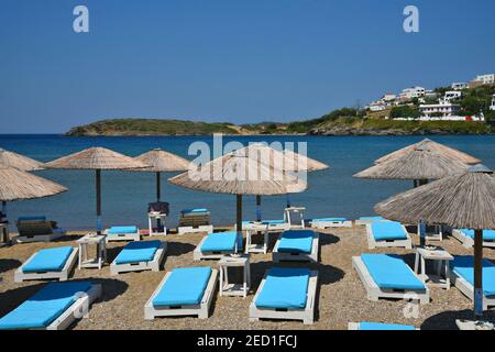 Landschaft mit blauen langen Stühlen und Strohschirmen am Ufer des Hafens von Batsi auf der Insel Andros, Kykladen Griechenland. Stockfoto