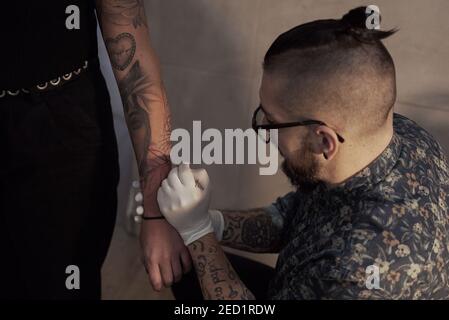 Hoher Winkel der männlichen Tätowierer macht Freihand-Tattoo und Zeichnung Skizze auf Arm der Ernte Client im Salon Stockfoto
