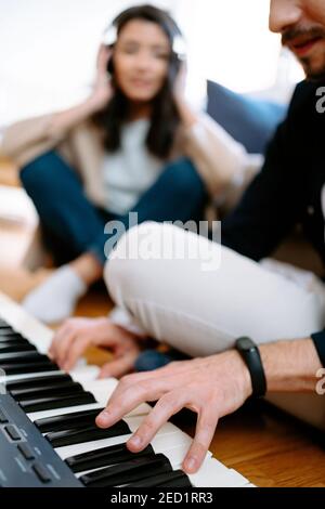 Mann spielt Synthesizer und Frau hört Musik in Kopfhörern, während sie zu Hause auf dem Boden sitzt und einen neuen Song aufnimmt Stockfoto