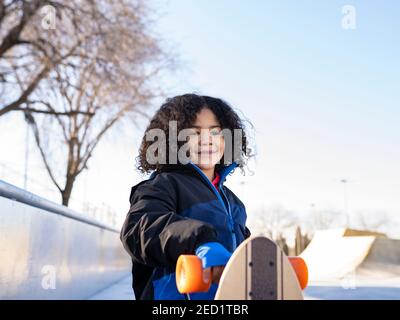 Entzücktes ethnisches Kind mit Afro-Frisur stehend mit Longboard auf Böschung an sonnigen Tag und Blick weg Stockfoto