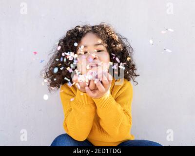 Fröhliche ethnische Kind mit lockigen Haaren weht bunten Papier Konfetti Bei der Kamera am sonnigen Tag gegen graue Wand Stockfoto