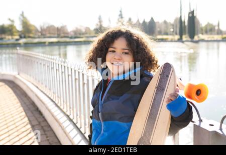 Entzücktes ethnisches Kind mit Afro-Frisur stehend mit Longboard auf Böschung an sonnigen Tagen und Blick auf die Kamera Stockfoto