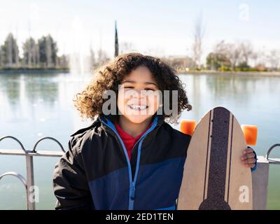 Entzücktes ethnisches Kind mit Afro-Frisur stehend mit Longboard auf Böschung an sonnigen Tagen und Blick auf die Kamera Stockfoto