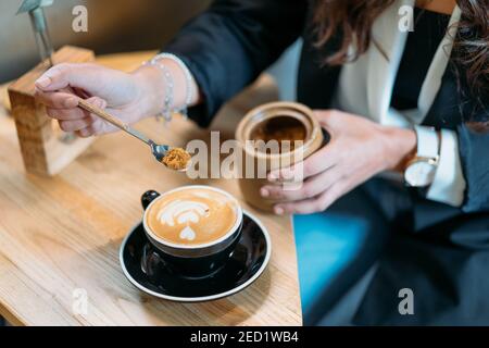 Von oben der Ernte anonyme Unternehmerin in stilvollem Outfit Hinzufügen Löffel braunen Zucker in Tasse mit Cappuccino, während Kaffeepause in CAF Stockfoto