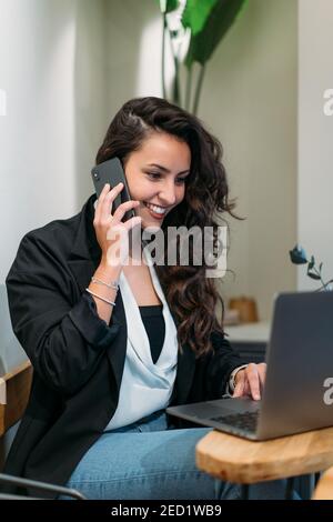 Glückliche Unternehmerin sitzt am Tisch und spricht auf dem Handy Beim Surfen Laptop und der Arbeit an Projekt Remote in Café Stockfoto