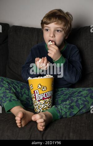 Staunender kleiner Junge sitzt bequem auf dem Sofa mit Popcornkübel Beim Essen und Fernsehen Stockfoto