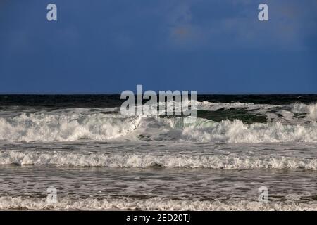 WINTERSZENE DUNKELBLAUER HIMMEL UND GRÜNE WELLEN MIT SCHAUM UND SPRAY KRACHEND AUF EINEN STRAND Stockfoto