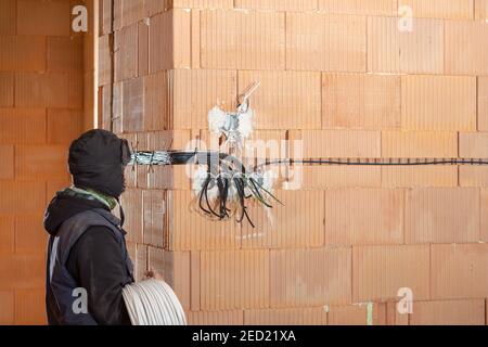 Ein Elektriker bei der Arbeit ist bereit, Steckdosen in unfertigen Haus aus Lehmziegeln gebaut zu installieren. Arbeiter bereit für die Verkabelung von Kabeln im Haus und Stockfoto