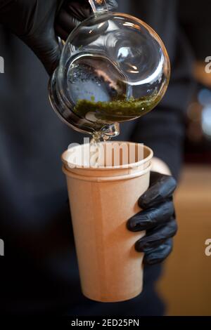 Nahaufnahme der Barista Hände, die heißen grünen Tee aus Glas eingiessen Teekanne in Papiertasse Stockfoto