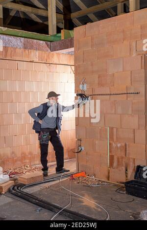 Ein Elektriker bei der Arbeit ist bereit, Steckdosen in unfertigen Haus aus Lehmziegeln gebaut zu installieren. Arbeiter bereit für die Verkabelung von Kabeln im Haus und Stockfoto