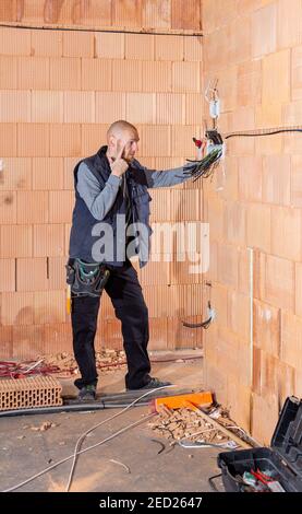 Ein Elektriker bei der Arbeit denken, was anfangen zu installieren zuerst in Steckdosen in unfertigen Haus aus Lehmziegeln gebaut. Arbeiter bereit für die Verdrahtung von Kabeln Stockfoto