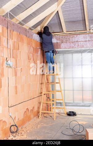 Elektriker, der auf einer Leiter steht und Kabel in den Kanal / Nut zusammen mit elektrischen Boxen in unfertigen Haus aus Lehmziegeln gebaut installiert. W Stockfoto