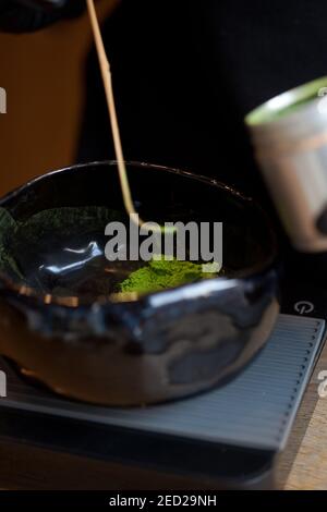 Wiegen von grünem Pulver Matcha Tee in dunkler Keramik-Schüssel auf Waage, Makro Nahaufnahme, keine Menschen Stockfoto