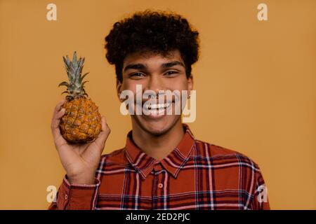 Foto von niedlichen afrikanischen männlichen Landwirt hält Ananas und Schleim. Der Mann trägt kariertes Hemd, isolierter brauner Hintergrund Stockfoto