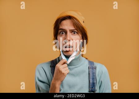 Foto von niedlichen weiblichen Landwirt hält Gesichtsmaske für Quarantäne Zeit macht überrascht Gesicht. Trägt Jeansüberkleidung und Hut, isolierte braune Farbe Hintergrund Stockfoto