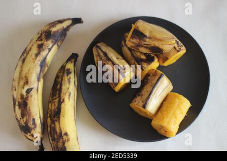 Reife Kochbanane gedämpft und auf dunkelgrauem mittelgroßem Teller serviert. Aufnahme auf weißem Hintergrund Stockfoto