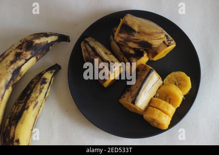 Reife Kochbanane gedämpft und auf dunkelgrauem mittelgroßem Teller serviert. Aufnahme auf weißem Hintergrund Stockfoto