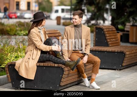 Glückliche junge stilvolle Frau sitzt auf Holzbank neben Ihr Freund, während beide plaudern während chill im Park Am Wochenende Stockfoto