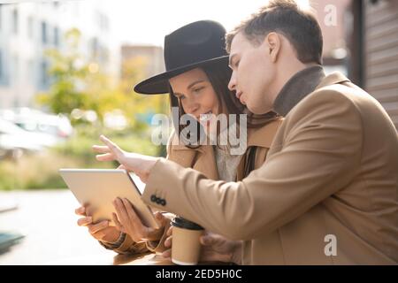 Junges Paar in stilvollen Casualwear Ruhe im Freien Café und Mit Diskussion über Online-Informationen, während Kerl zeigt auf Tablet Anzeigen Stockfoto