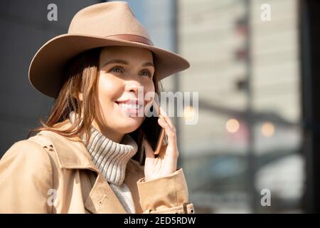 Happy Young wunderschöne Brünette weiblich in Hut, beige Trenchcoat und grauen Pullover im Gespräch auf Handy in städtischen Umgebung Stockfoto